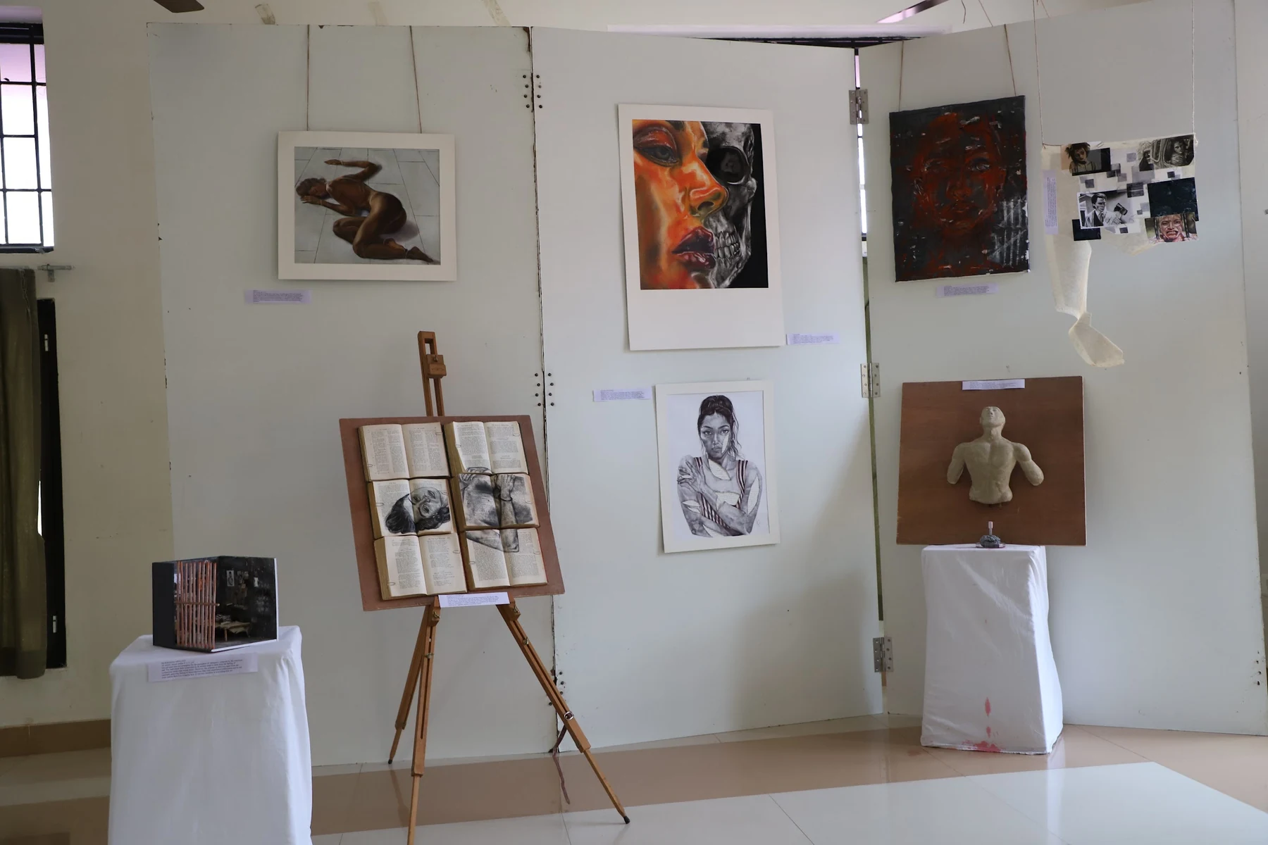 A display wall of drawings and paintings, with an easel holding a drawing on book pages, a relief torso and a boxed model on plinths