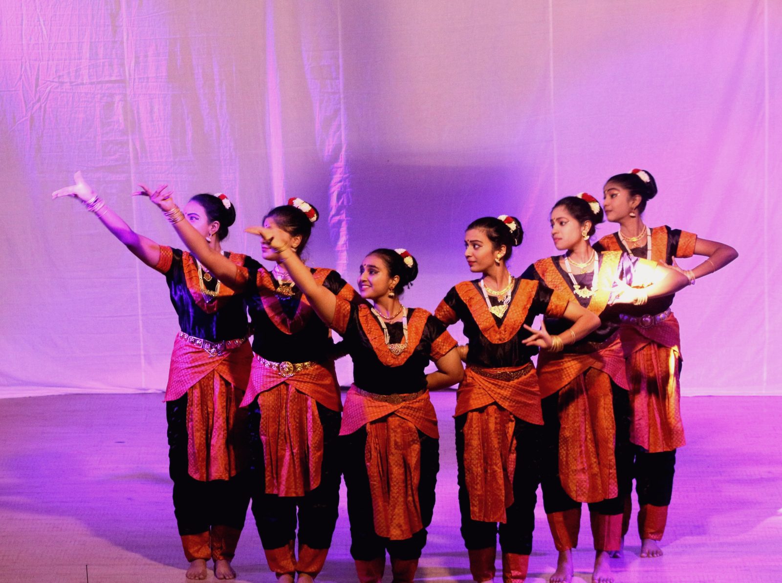 A row of dancers in costume raising their arms on a stage lit in violet