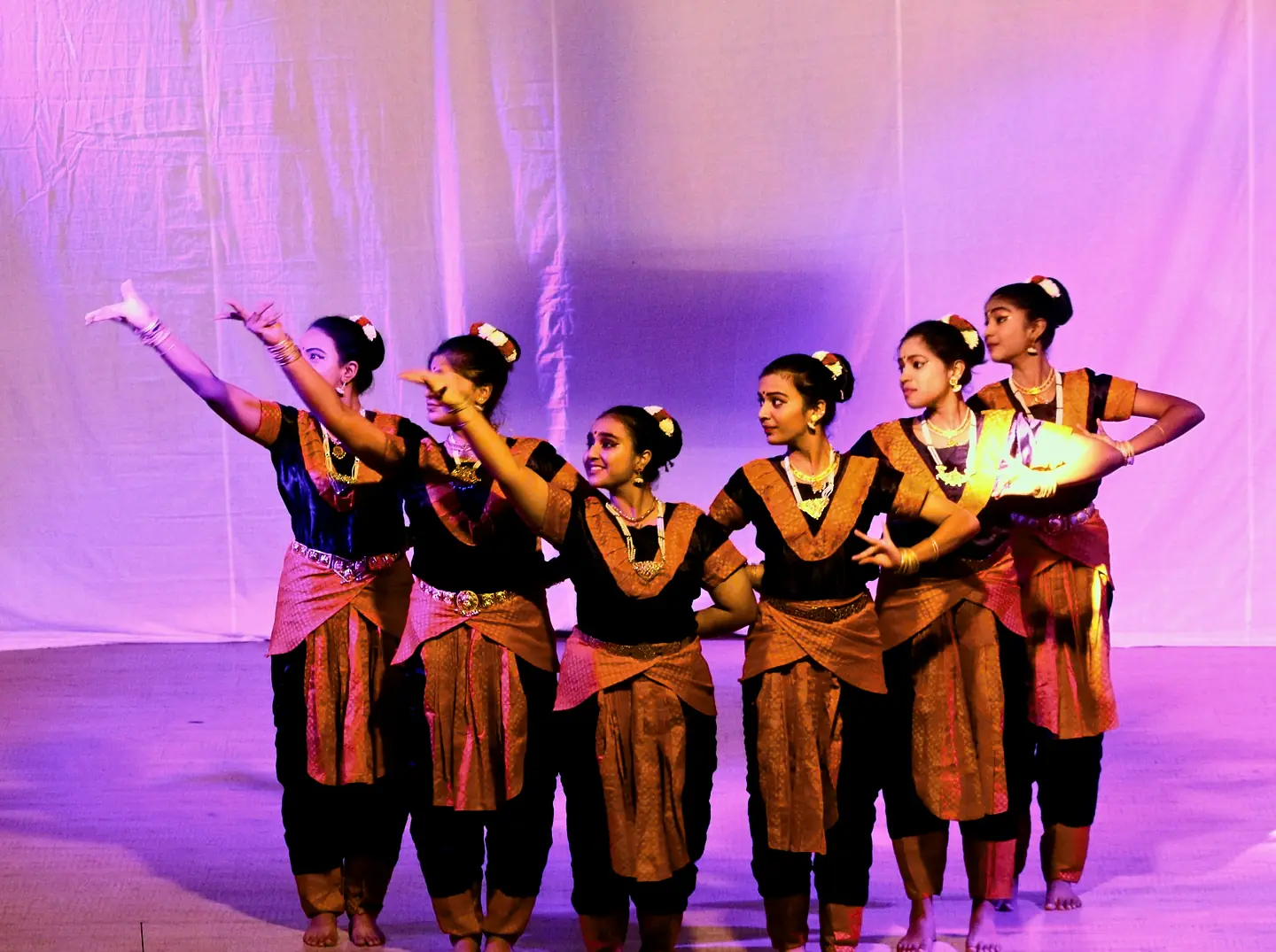A row of dancers in costume raising their arms on a stage lit in violet