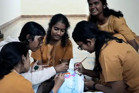 Four students bent over a pale blue clay pot, painting a band of pink and orange with fine brushes