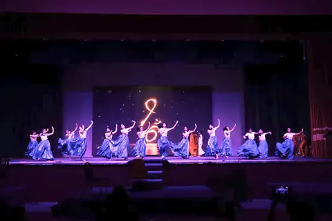 Dancers in blue and purple costumes on a stage lit in violet, with a glowing symbol at the back