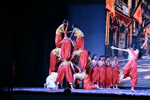 Dancers in red and gold costumes on stage, some of them stacked in a human pyramid, in front of a screen showing a street of garlanded doorways