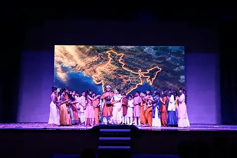 A large group of students in white and coloured clothes on stage, in front of a screen showing a glowing outline map of India