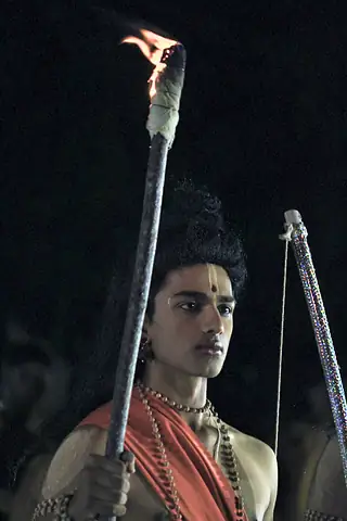 A student in costume as Rama holding a bow beside a burning torch