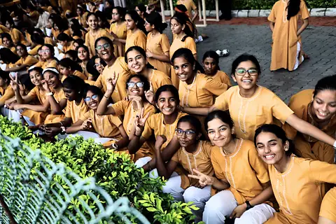 Girls in saffron kurtas seated close together on the bank, cheering