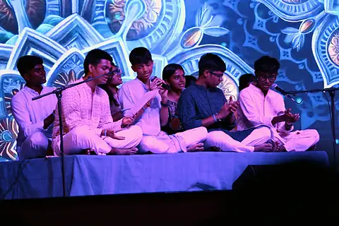 A group of students seated on stage singing before a blue projected mandala