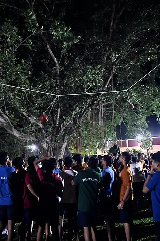 A crowd of students under a tree at night, looking up at a pot on a rope