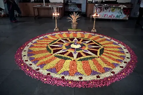 A large flower pookalam on the dining-hall floor between two lit brass lamps