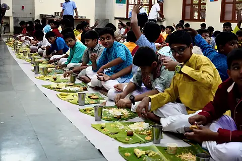 A long row of students seated on the floor eating the sadhya from banana leaves