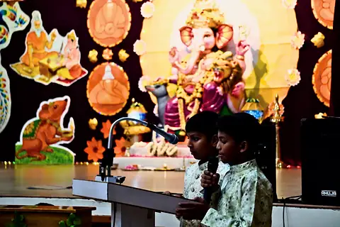 Two boys at a lectern before a decorated Ganesh backdrop