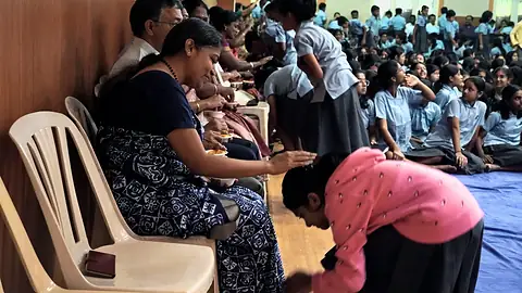 A woman seated on a chair as a student in pink bends to touch her feet, with rows of students seated behind