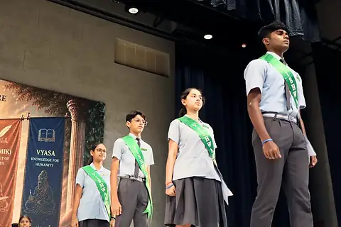 Three students wearing green sashes on a stage before a Vyasa banner