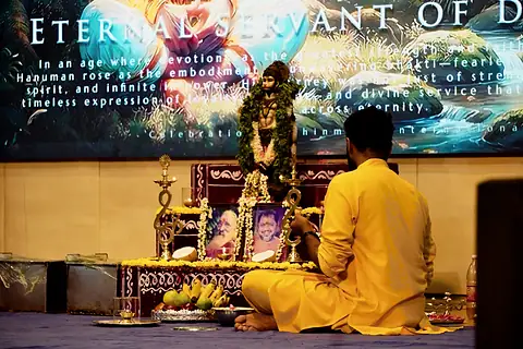 A student in a yellow kurta seated before a garlanded murti, lamps and offerings