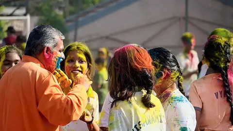 A man in an orange kurta smearing colour on a smiling student among others with coloured faces at Holi