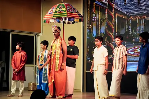 Students on a stage in a skit, one standing beneath a colourful ceremonial umbrella