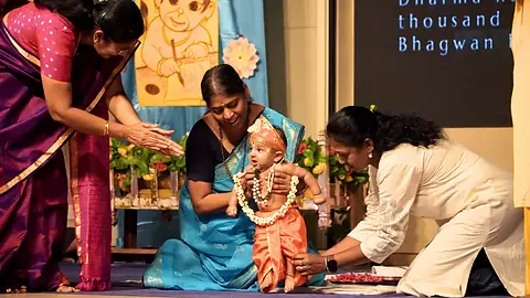 A garlanded child dressed as Bal Ram seated at a decorated altar as women make offerings