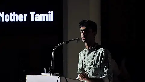 A student speaking at a microphone beneath a sign that reads Mother Tamil