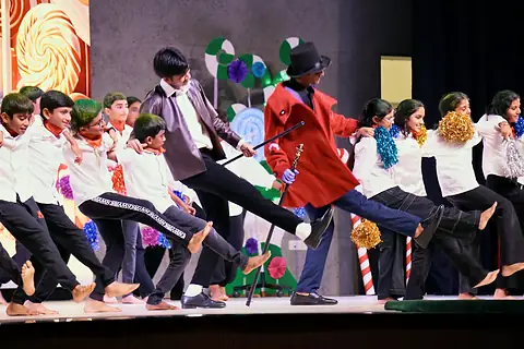 The company dancing across the stage before a candy-land backdrop, a lead in a red coat and top hat among them