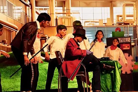 Actors in a factory set, one in a red coat seated with a cane