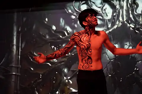 An actor in red swirling body paint, arms spread, against a dark backdrop