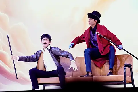 Two actors on a sofa before a white-and-peach backdrop, one in a red coat holding a cane