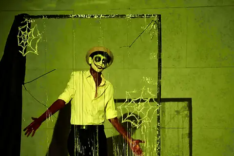 An actor in skull face paint in a green set strung with cobwebs