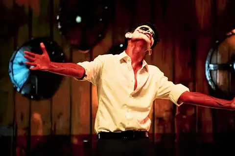 An actor in skull face paint with red-painted hands, arms spread, before a wooden wall hung with shields