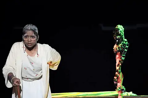 An actor made up with grey hair, leaning on a stick in the dark