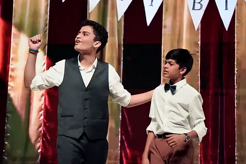 An actor in a waistcoat with a fist raised beside a younger actor in a bow tie, bunting above