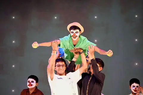 A performer in clown face paint lifted high by others