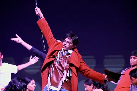 An actor in a red coat, arm raised, among dancers under purple light