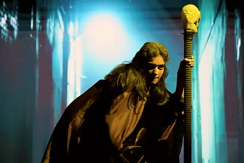 A figure in a shaggy cloak holding a skull-topped staff, lit green and gold