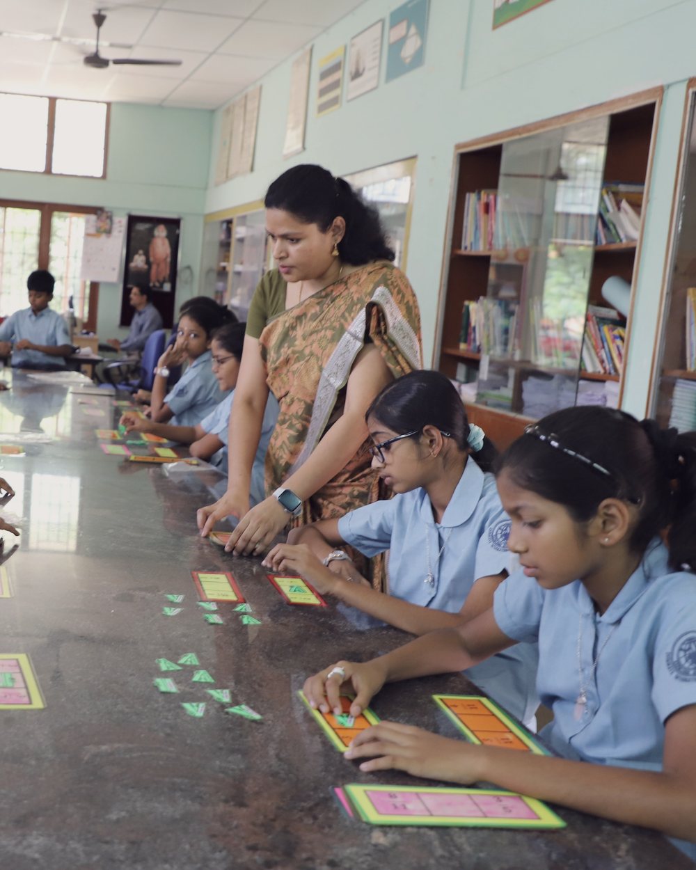 Junior School students at a long classroom bench, working with coloured cards while their teacher looks on