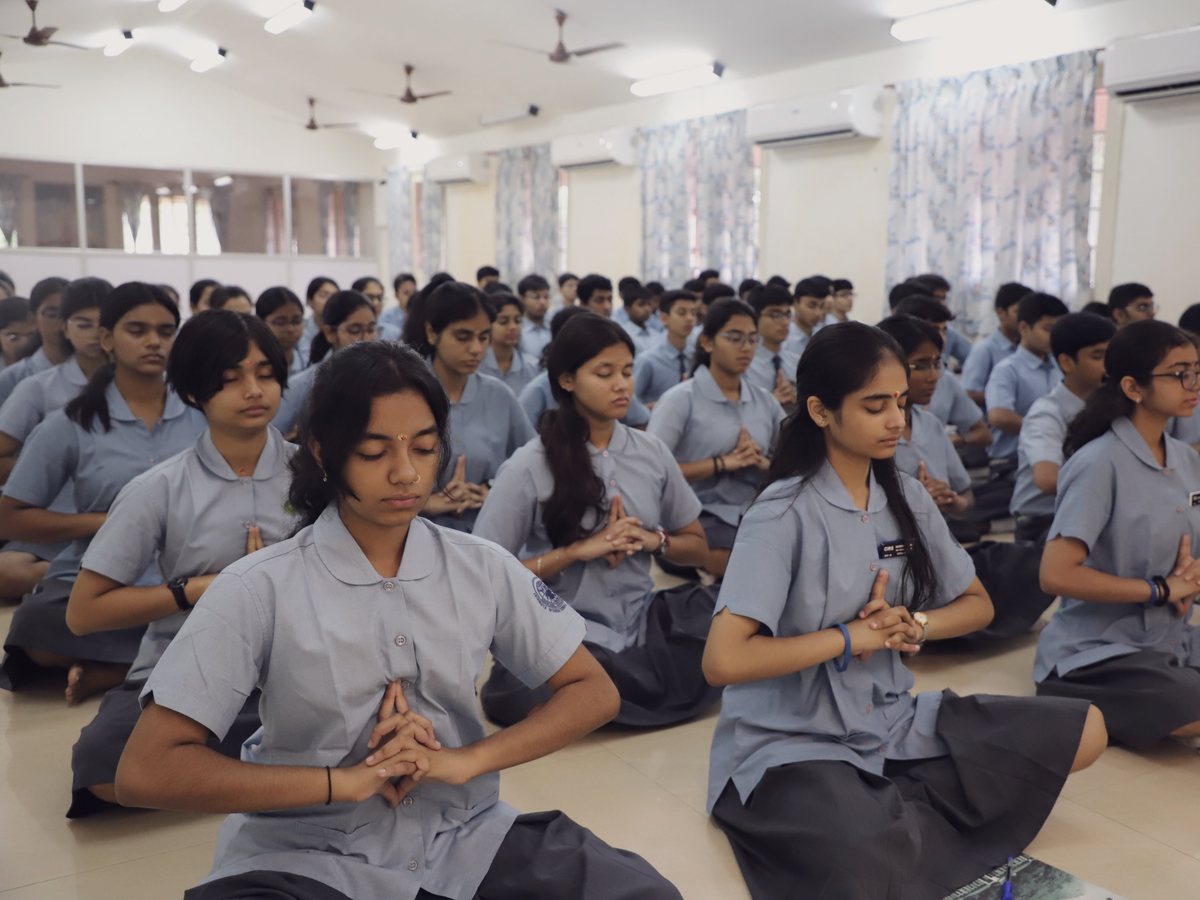 Students seated in rows in a hall, eyes closed and palms joined