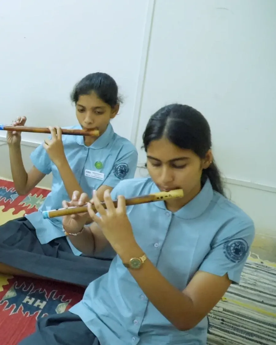 Two students in school uniform seated on a rug, playing bamboo flutes