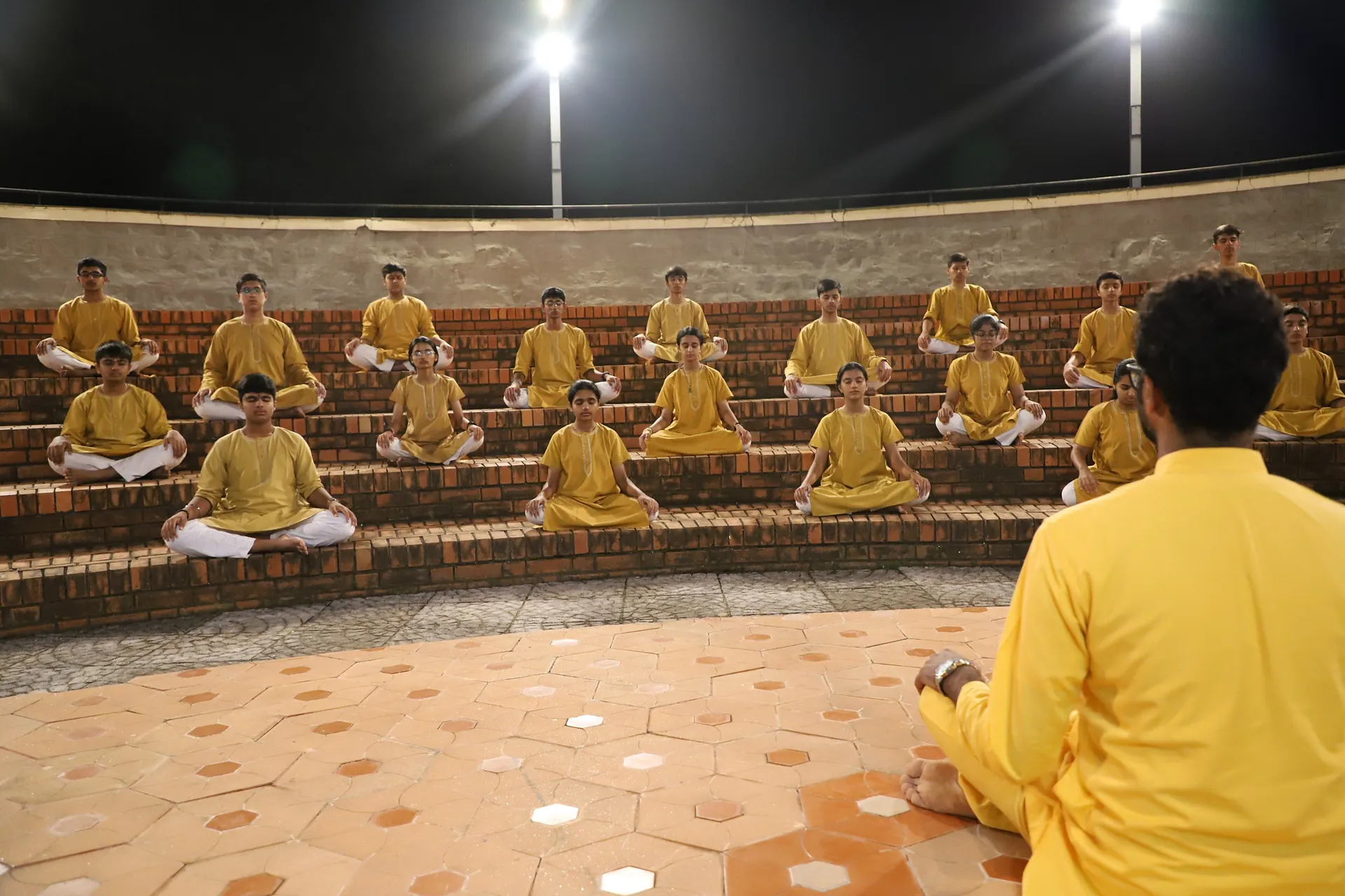 Students in yellow kurtas seated in the lit amphitheatre after dark, a teacher facing them