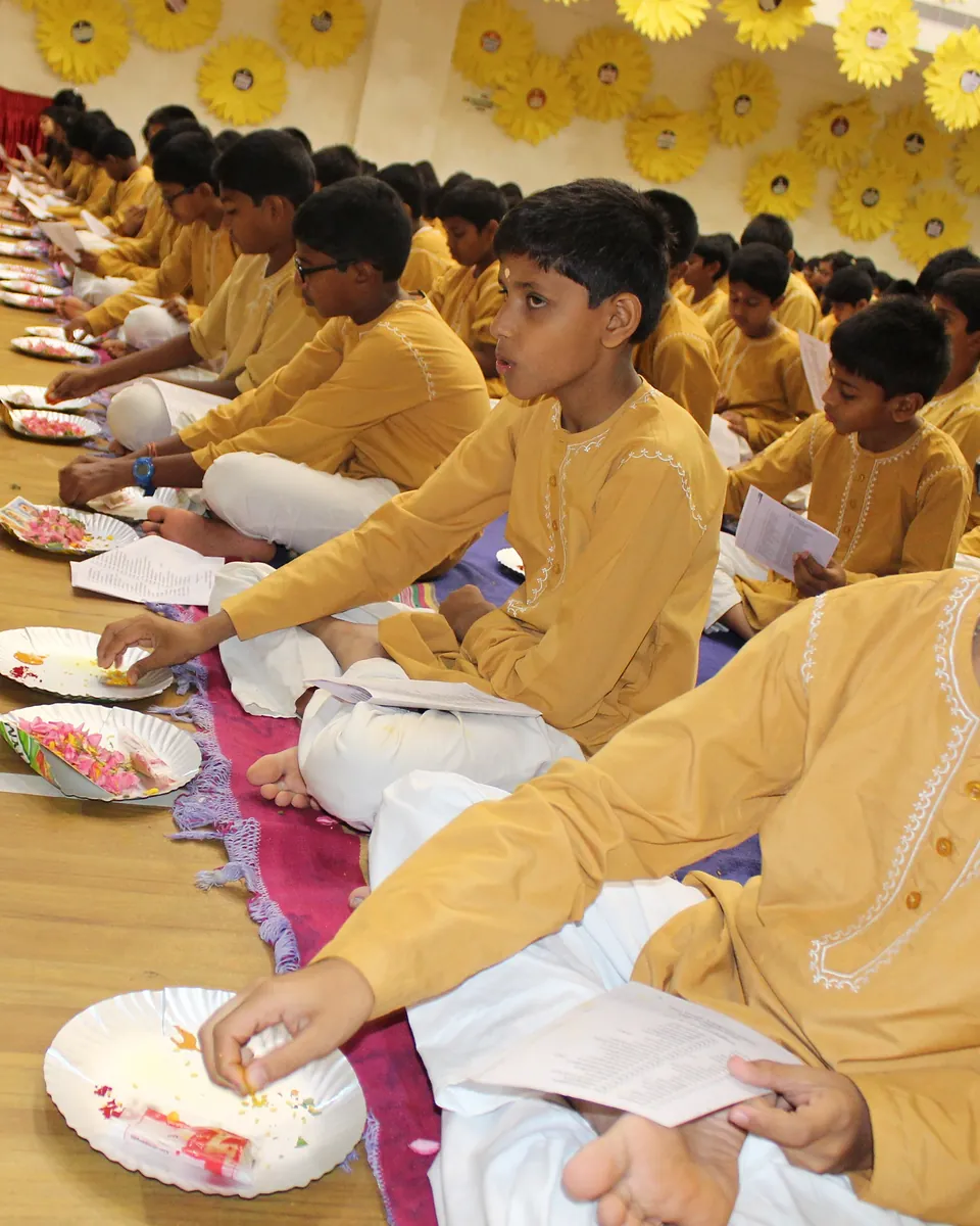 Students in yellow kurtas seated in a long row on the floor, each with a plate of flowers for a puja