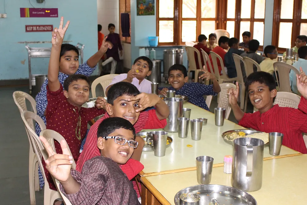 Junior boys at a dining-hall table with steel plates and tumblers, waving at the camera
