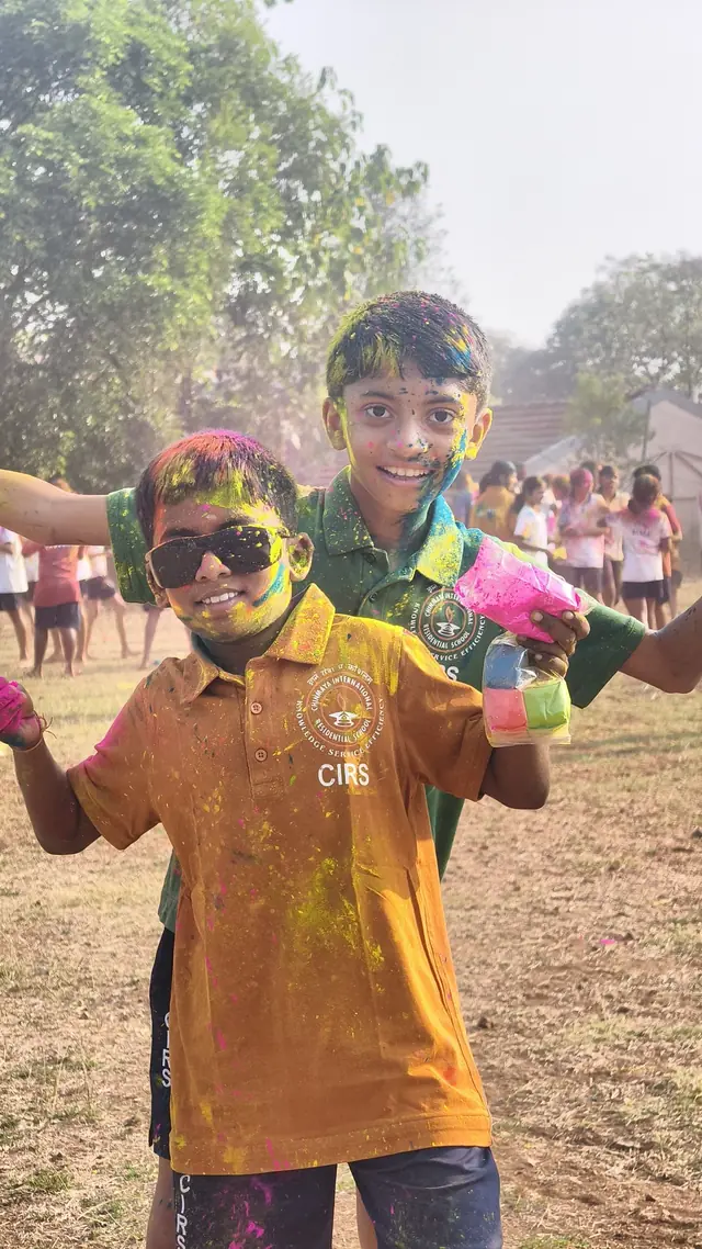 A boy in an orange CIRS shirt covered in colour, grinning, a friend behind him