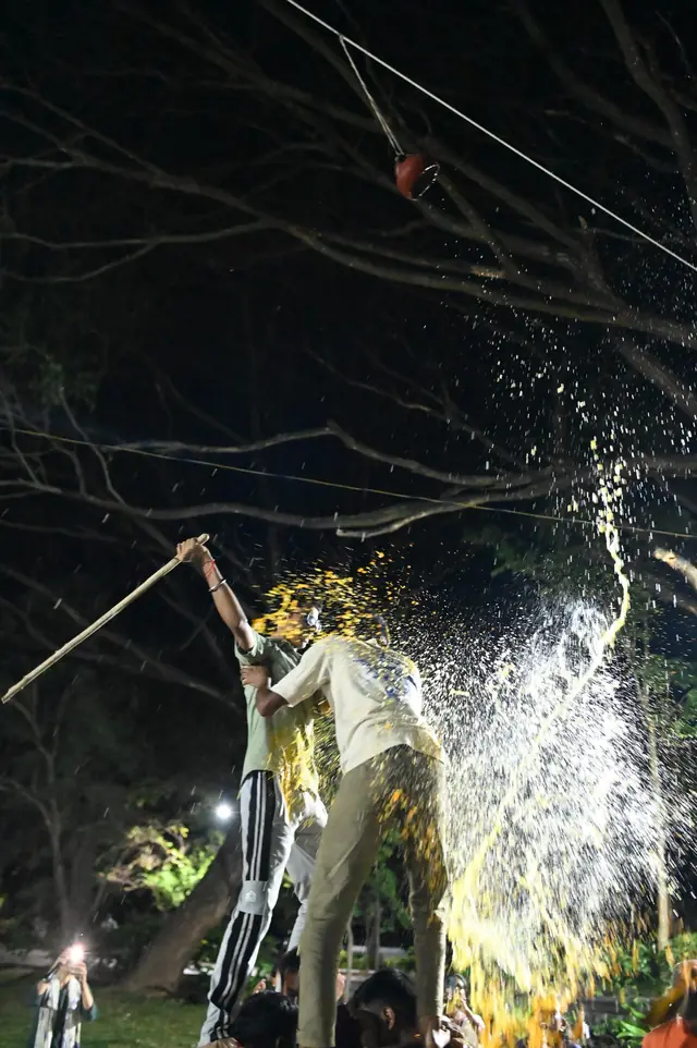A student at the top of a human pyramid strikes the pot, and its contents burst out in the floodlight