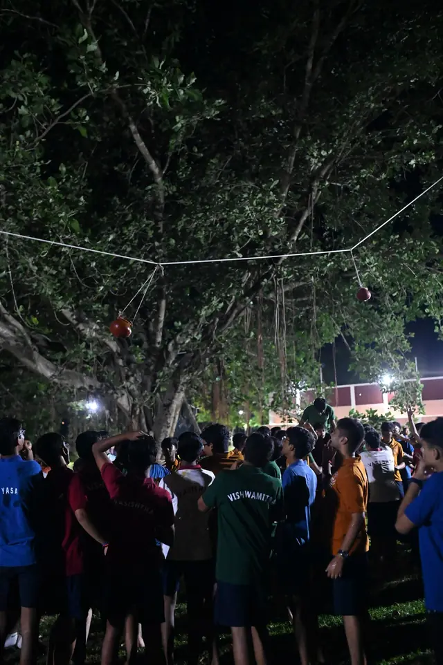 A crowd of students under a tree at night, looking up at a pot on a rope