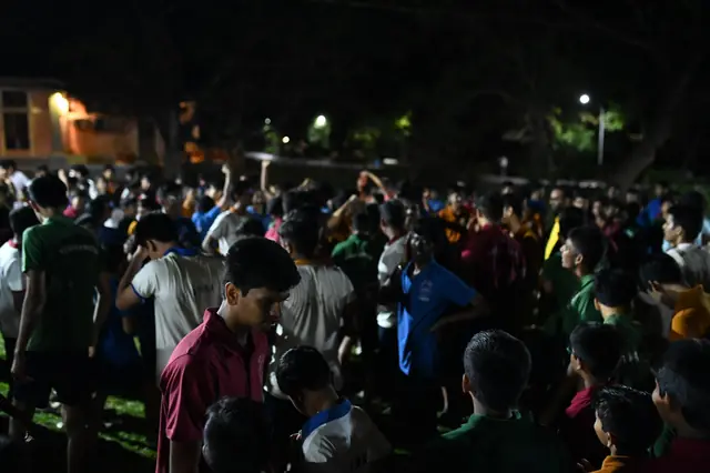 Students crowded on a lawn at night under the trees and lamps