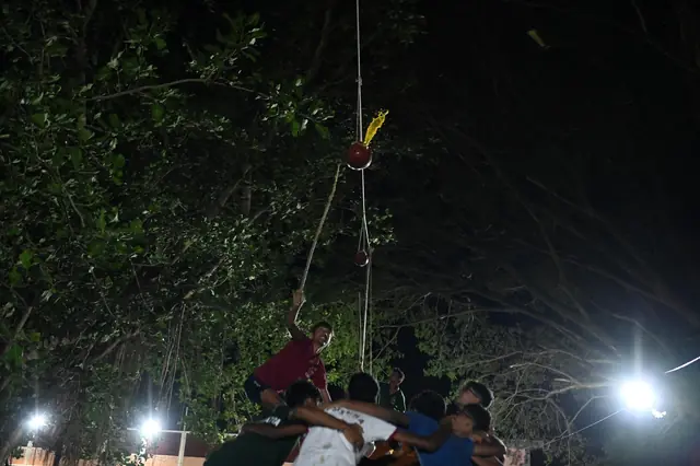 Students building a human pyramid under a tree at night, a pot hanging above them