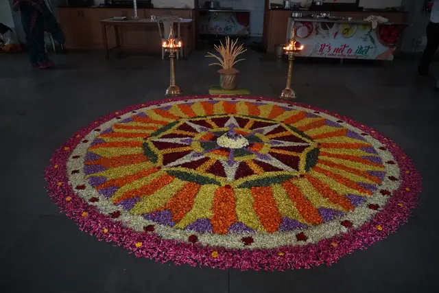 A large flower pookalam on the dining-hall floor between two lit brass lamps