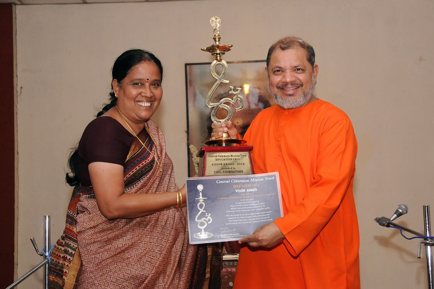 A trophy inscribed Central Chinmaya Mission Trust Education Cell Vision Award 2012, held up with its certificate by a woman in a sari and a swami in saffron
