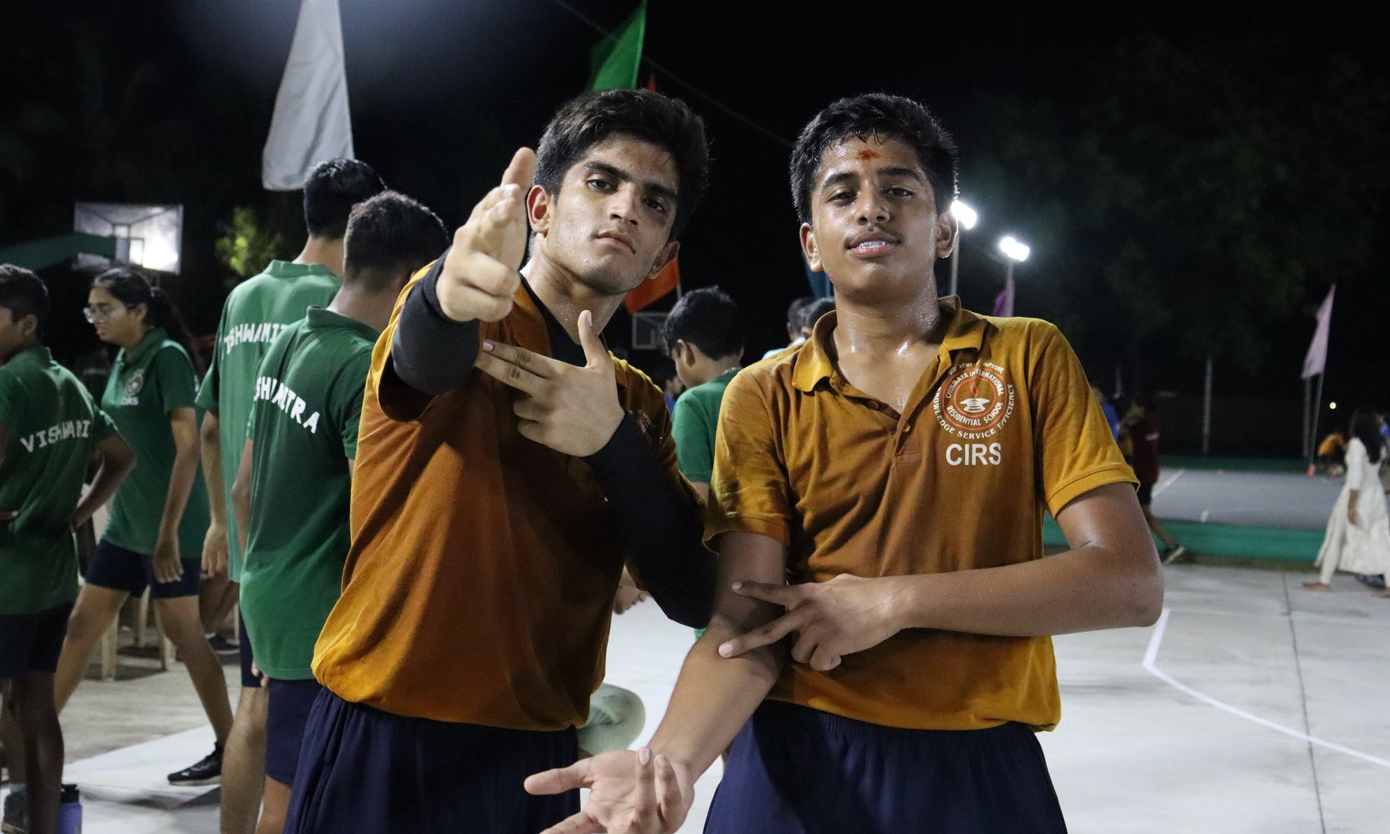 Two CIRS students pose together on the basketball court, with students in named green house shirts behind them