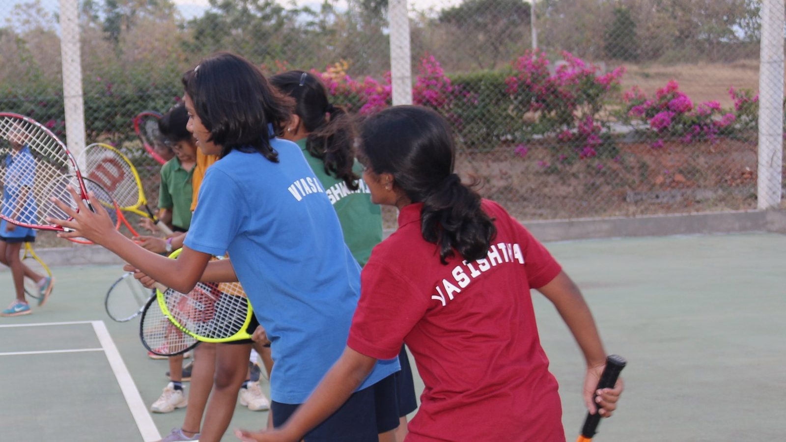 Students practising tennis, with Vyasa and Vasishta named on their blue and red shirts