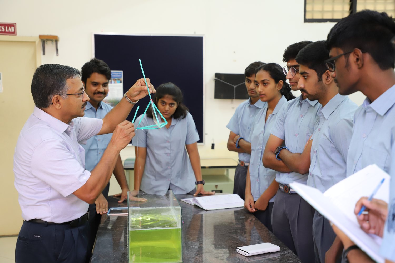 A teacher holding up a model over a tank of green water in a laboratory while students in uniform lean in to watch.