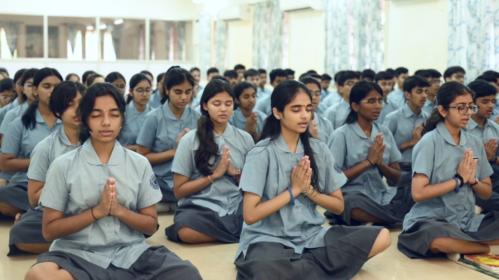 Rows of students in uniform seated cross-legged on the floor of a hall, eyes closed and palms joined.