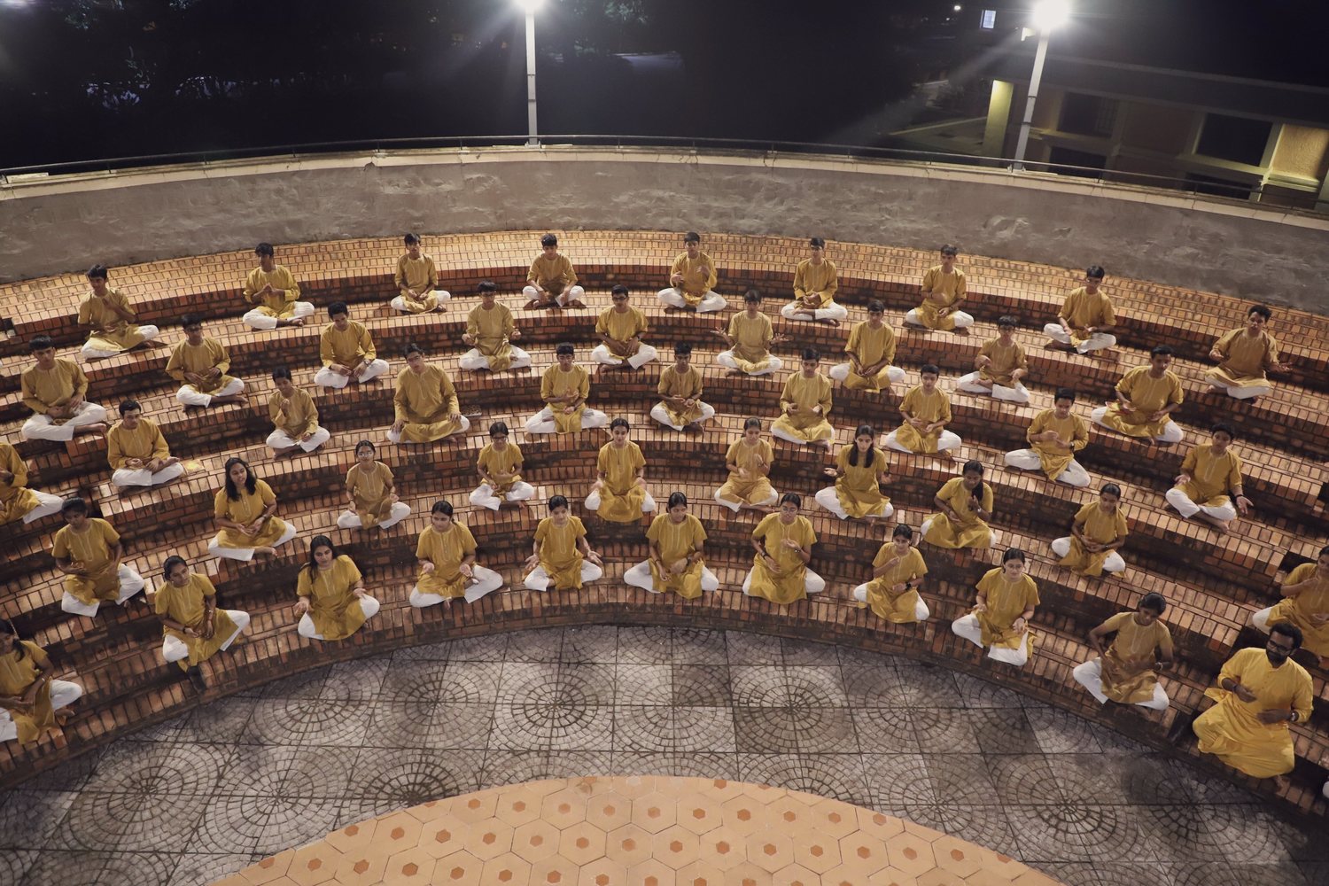 Students seated in the concentric brick rings of the CIRS amphitheatre after
                dark, under floodlights, during evening practice.